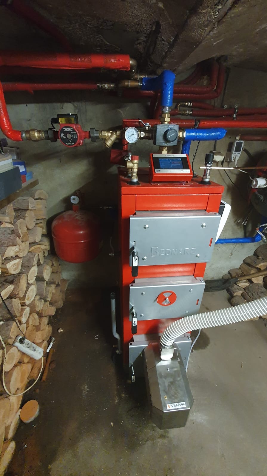Czerwony piec na paliwo stałe marki Bednarz z podajnikiem Venma, widoczne rury instalacji grzewczej, manometry, sterownik i zapas drewna obok.