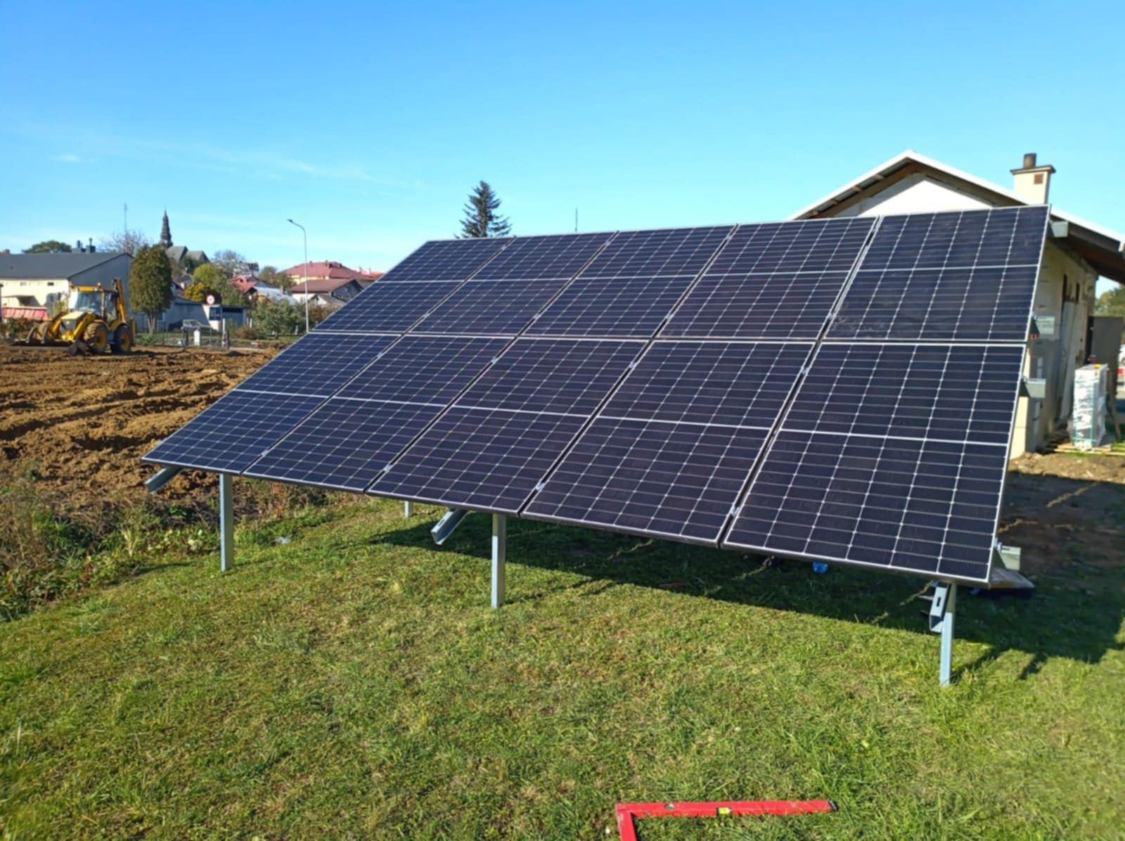 Panel fotowoltaiczny na metalowej konstrukcji, zamontowany na trawniku przed domem, z traktorem pracującym na polu w tle.
