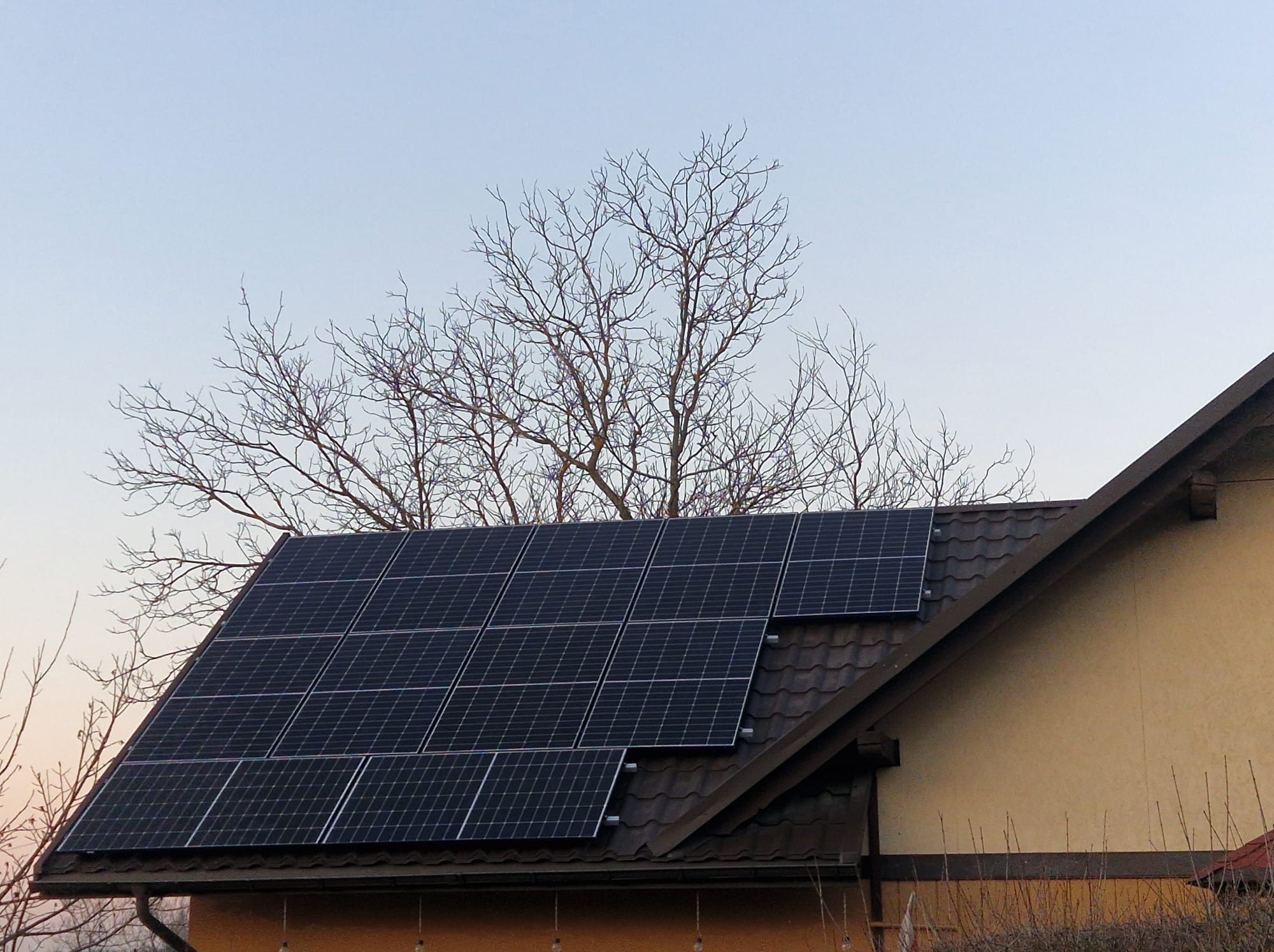 Panele słoneczne zamontowane na brązowym dachu domu jednorodzinnego, na tle błękitnego nieba i gałęzi drzewa bez liści.