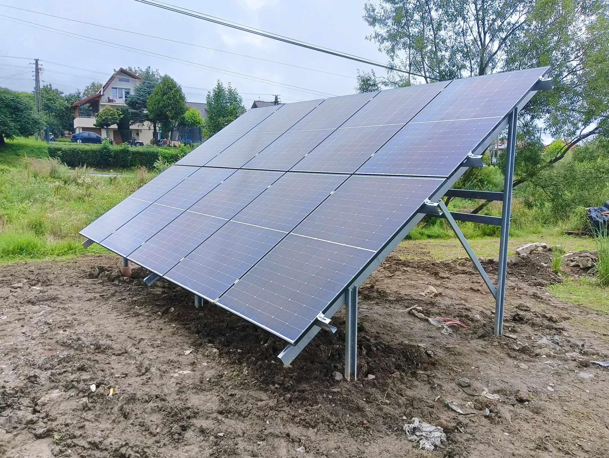 Panel słoneczny ustawiony na metalowej konstrukcji na ziemi, widoczny dom w tle, pochmurne niebo.