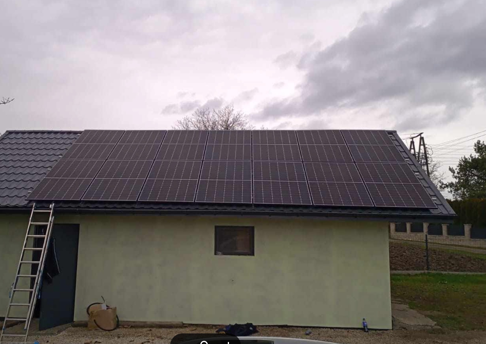 Panele słoneczne na dachu budynku gospodarczego z drabiną opartą o ścianę, widok z poziomu gruntu w pochmurny dzień.