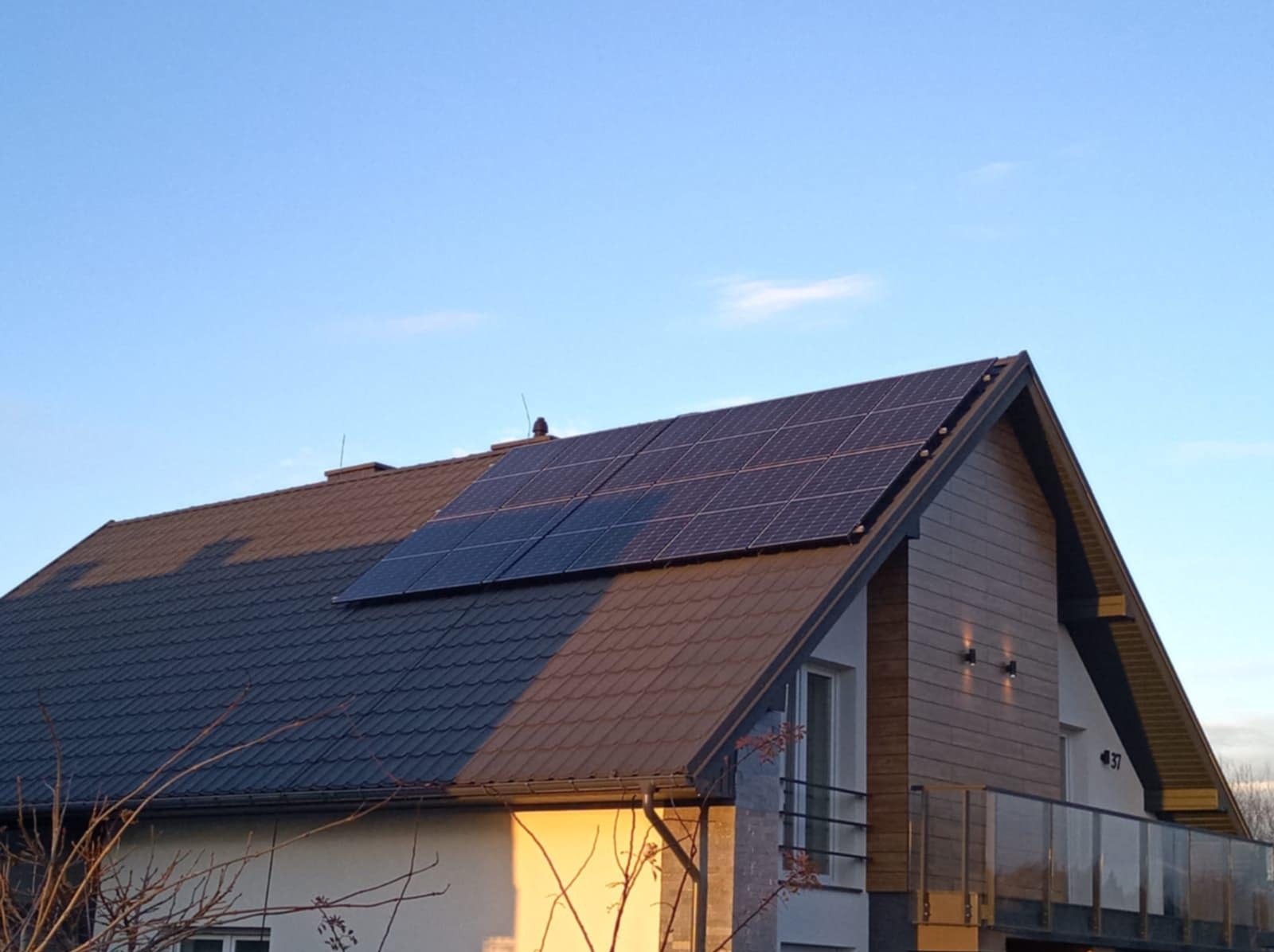 Panele fotowoltaiczne zamontowane na dachu dwuspadowym nowoczesnego domu z brązową dachówką i jasną elewacją w słoneczny dzień.