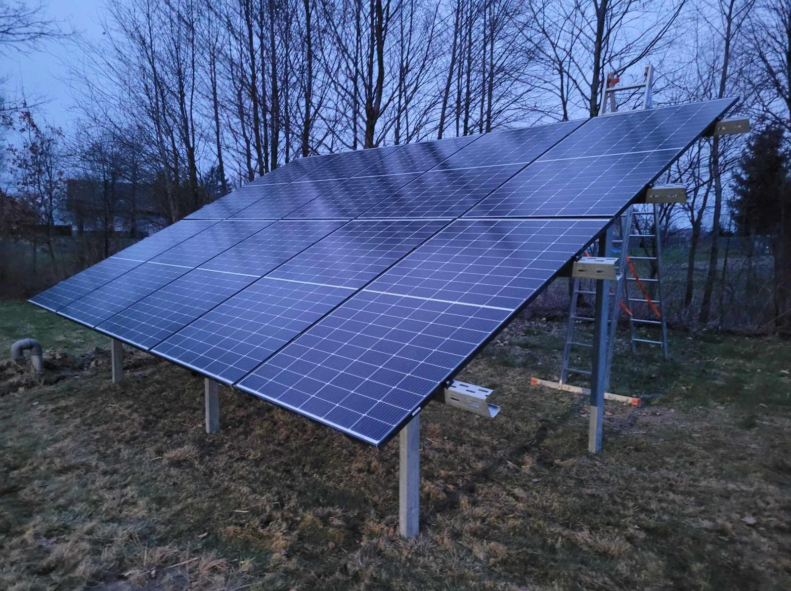 Panel słoneczny na betonowych słupach w pochmurny dzień, drabina oparta o konstrukcję, widoczne elementy montażowe.