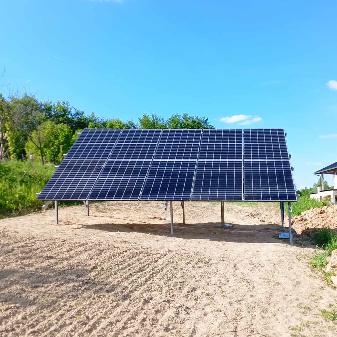 Panele słoneczne zamontowane na metalowej konstrukcji na piaszczystym terenie, widok z przodu, błękitne niebo w tle.