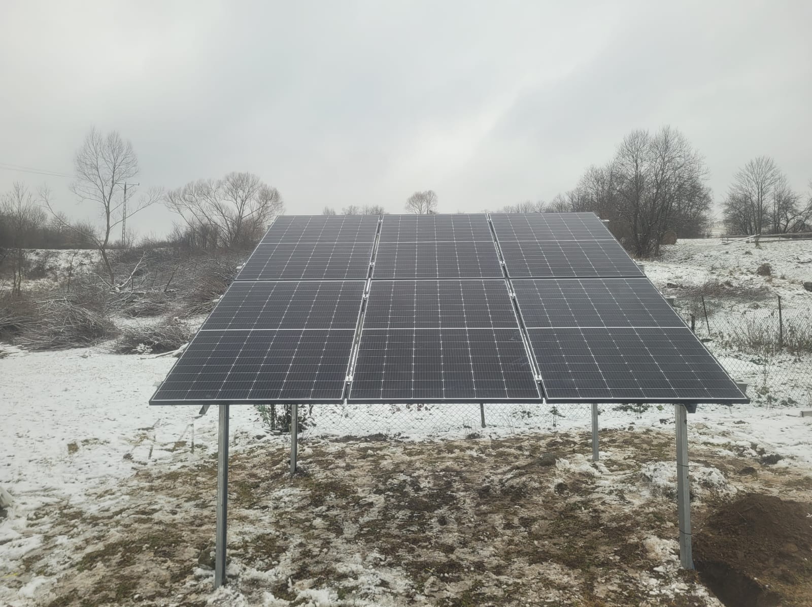 Panel słoneczny na metalowych podporach, częściowo pokryty śniegiem, na tle zimowego krajobrazu z drzewami bez liści i pochmurnym niebem.