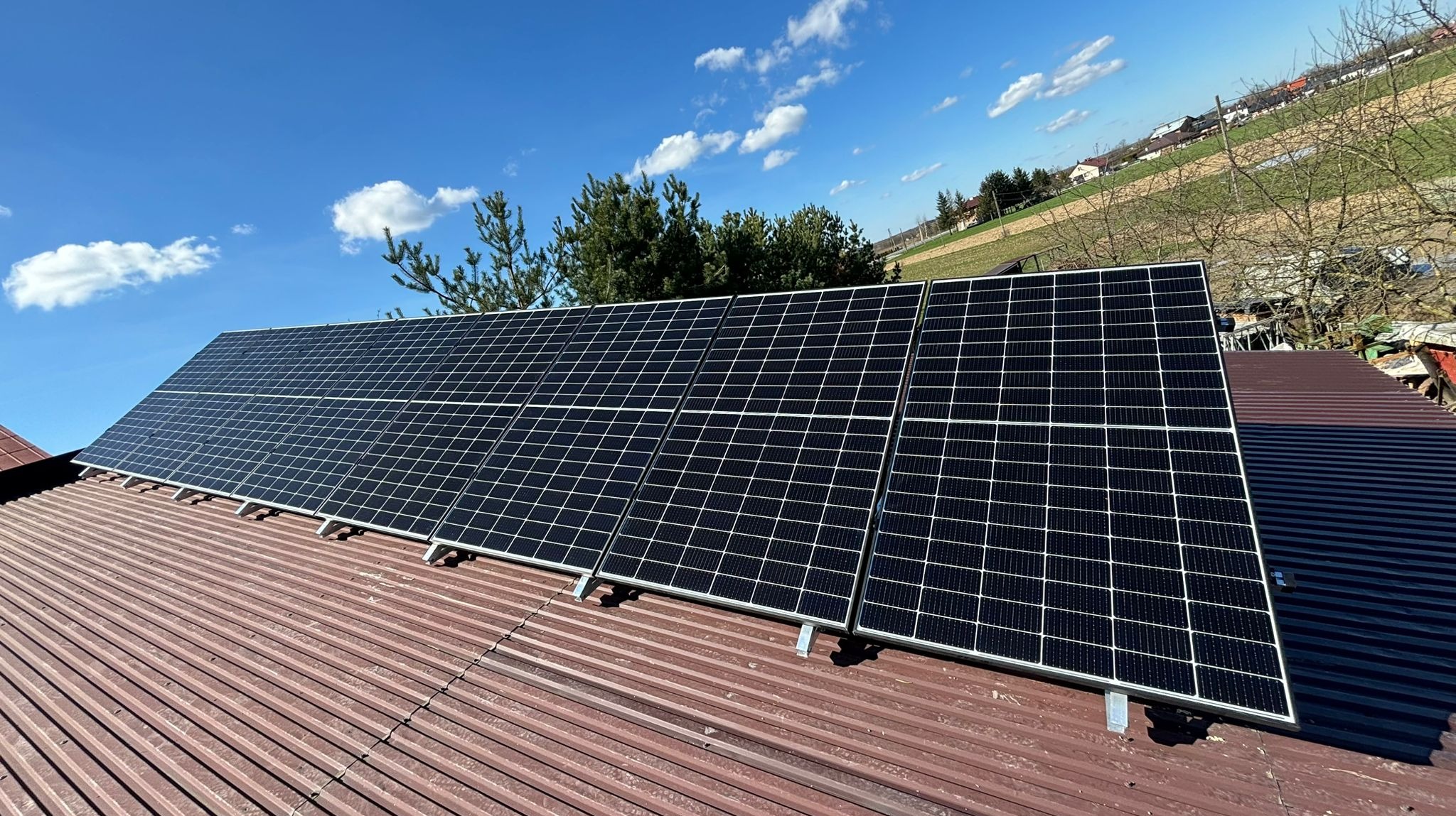 Panele słoneczne zamontowane na falistej, czerwonej blasze dachu, na tle błękitnego nieba z pojedynczymi obłokami i zielenią drzew w oddali.