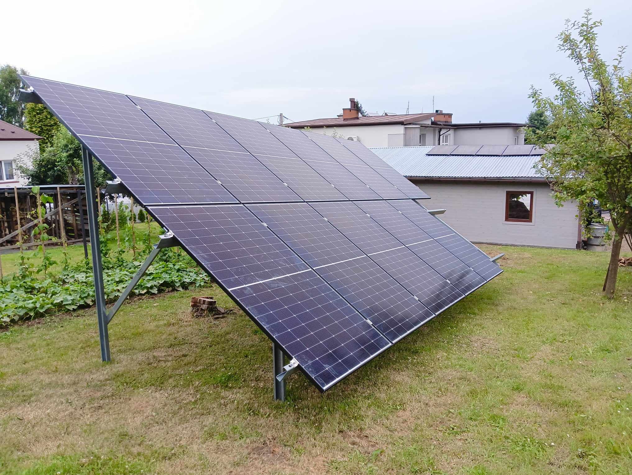 Zestaw paneli słonecznych ustawiony na trawniku w ogrodzie, z widocznymi budynkami mieszkalnymi w tle i fragmentem upraw warzywnych.