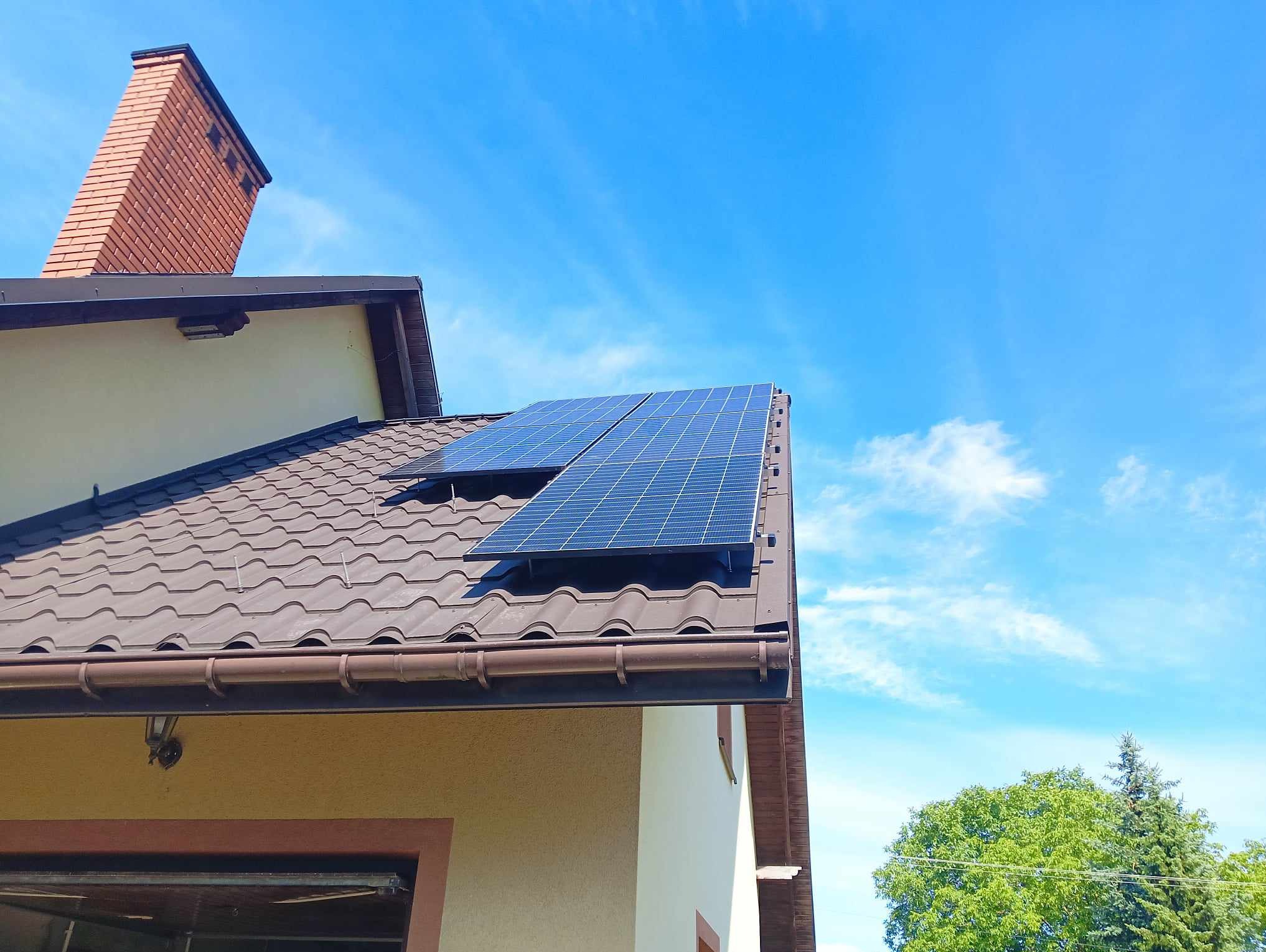 Dwa panele słoneczne na brązowym, skośnym dachu domu z ceglanym kominem, widoczne z dołu na tle błękitnego nieba z delikatnymi chmurami.
