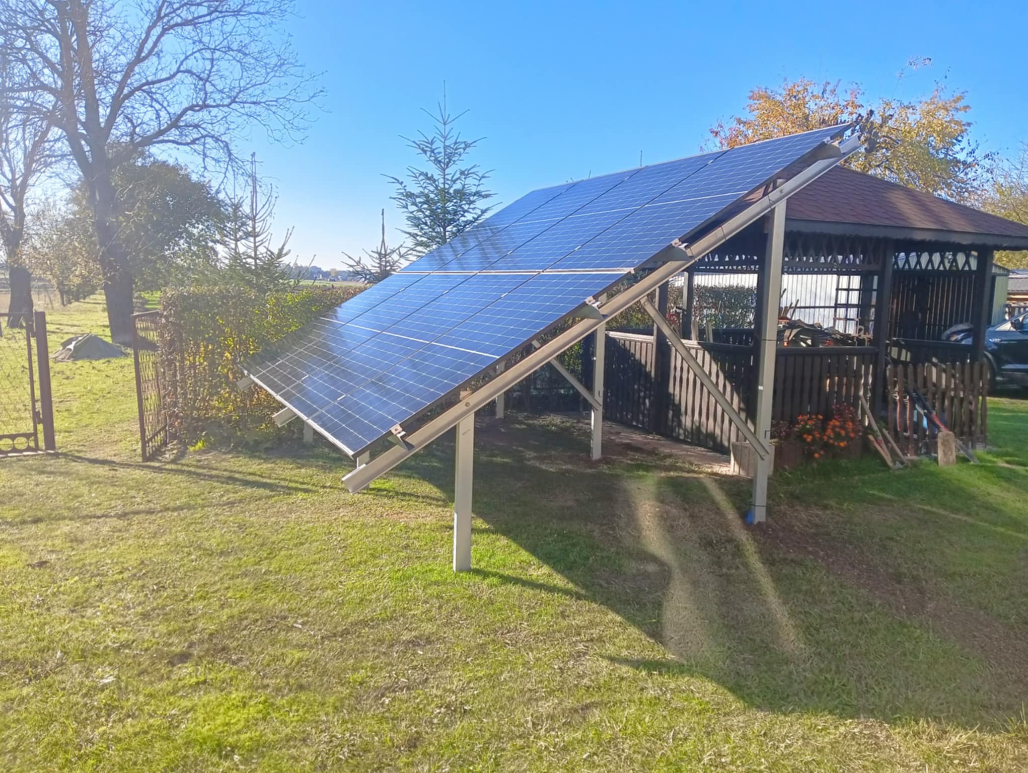 Instalacja fotowoltaiczna na trawniku obok altany z brązowym dachem w słoneczny dzień.