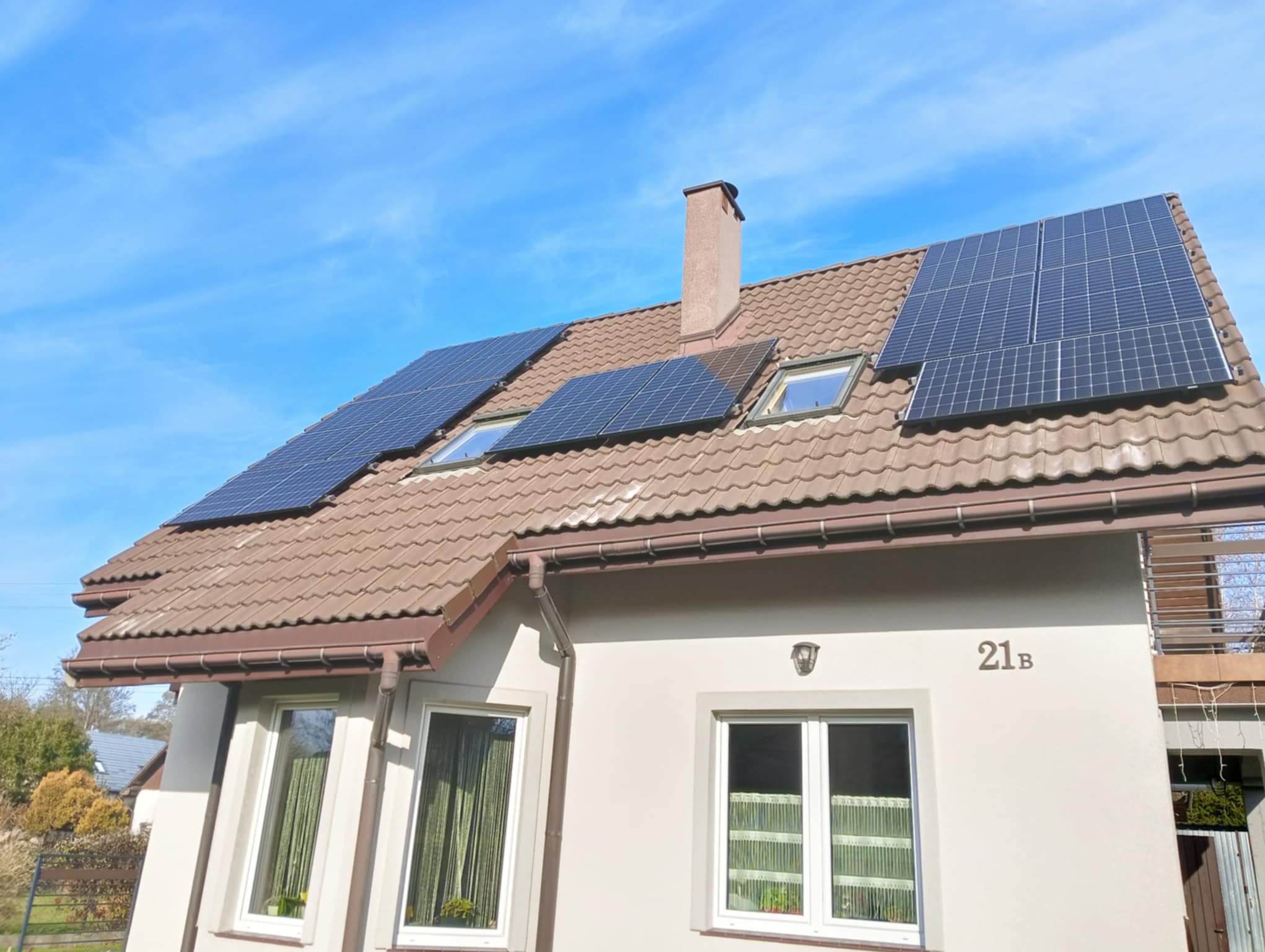 Dom jednorodzinny z brązową dachówką, na której zamontowano panele słoneczne, widoczne również okna dachowe i komin, na ścianie budynku numer 21b.