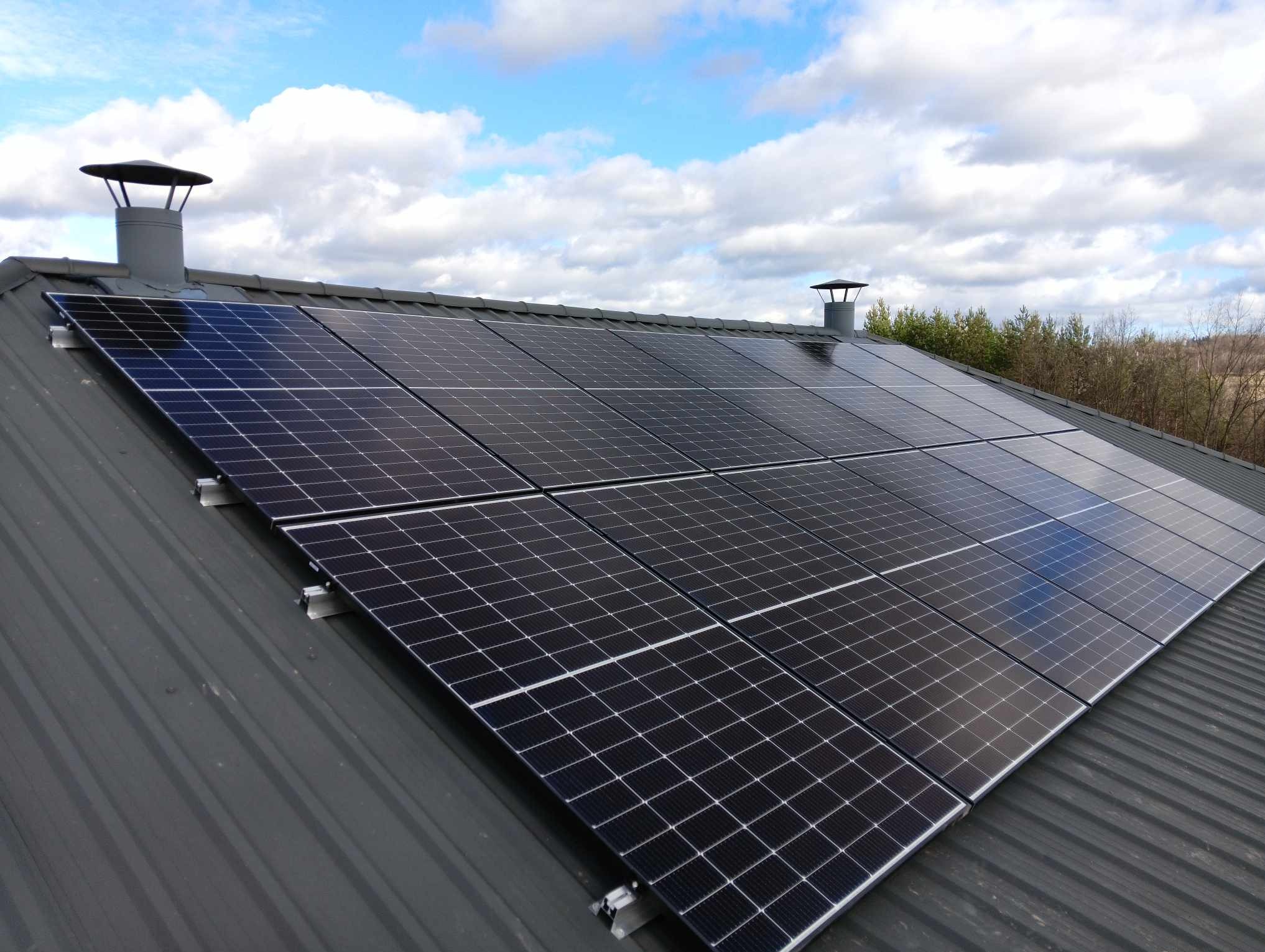 Instalacja paneli słonecznych na metalowym dachu domu w Rzeszowie, widoczne kominy wentylacyjne i błękitne niebo z chmurami odbijające się w panelach.