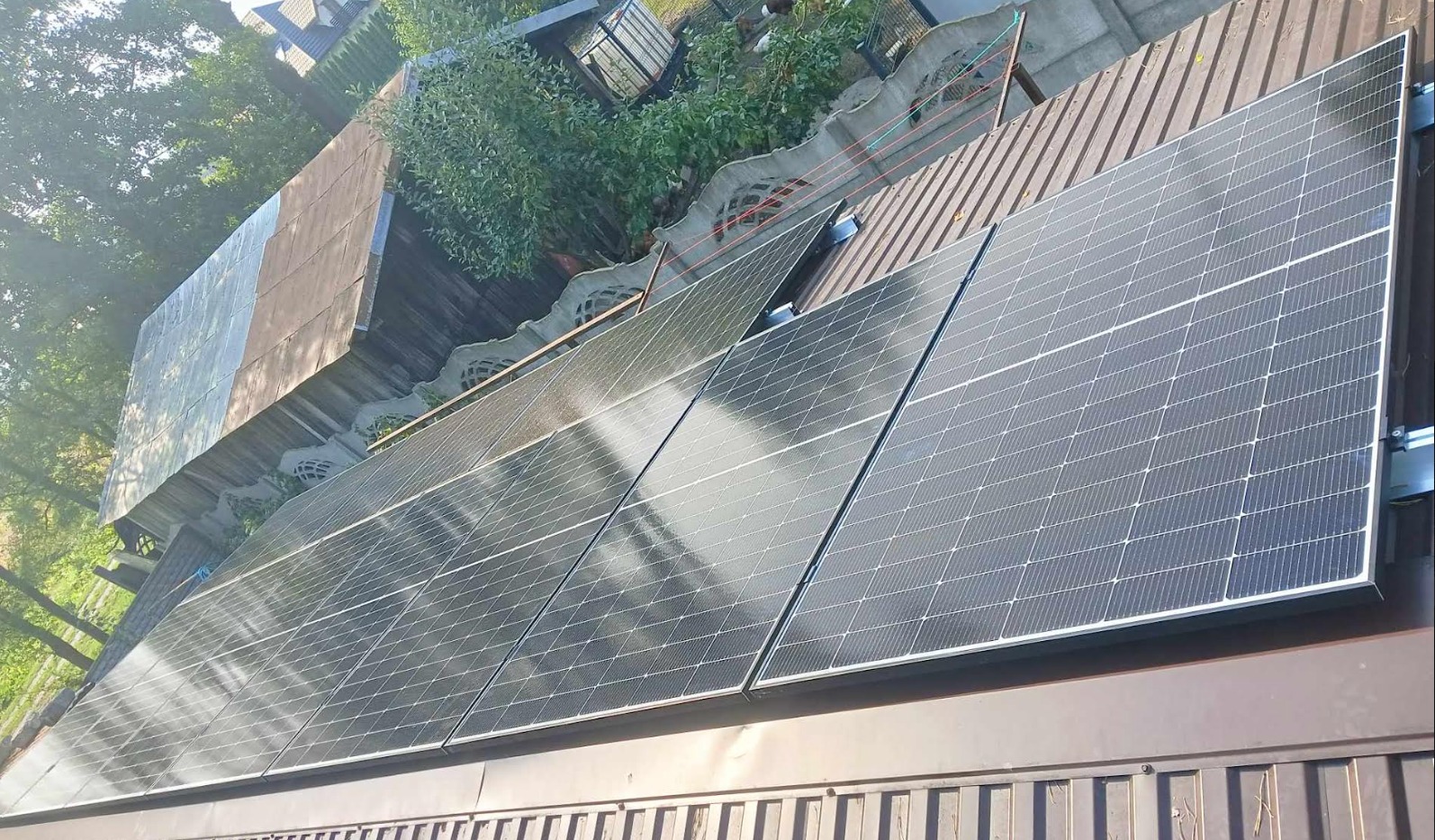 Panele słoneczne zamontowane na metalowym dachu, widoczne z perspektywy dachu sąsiedniego budynku, z zielenią drzew w tle.