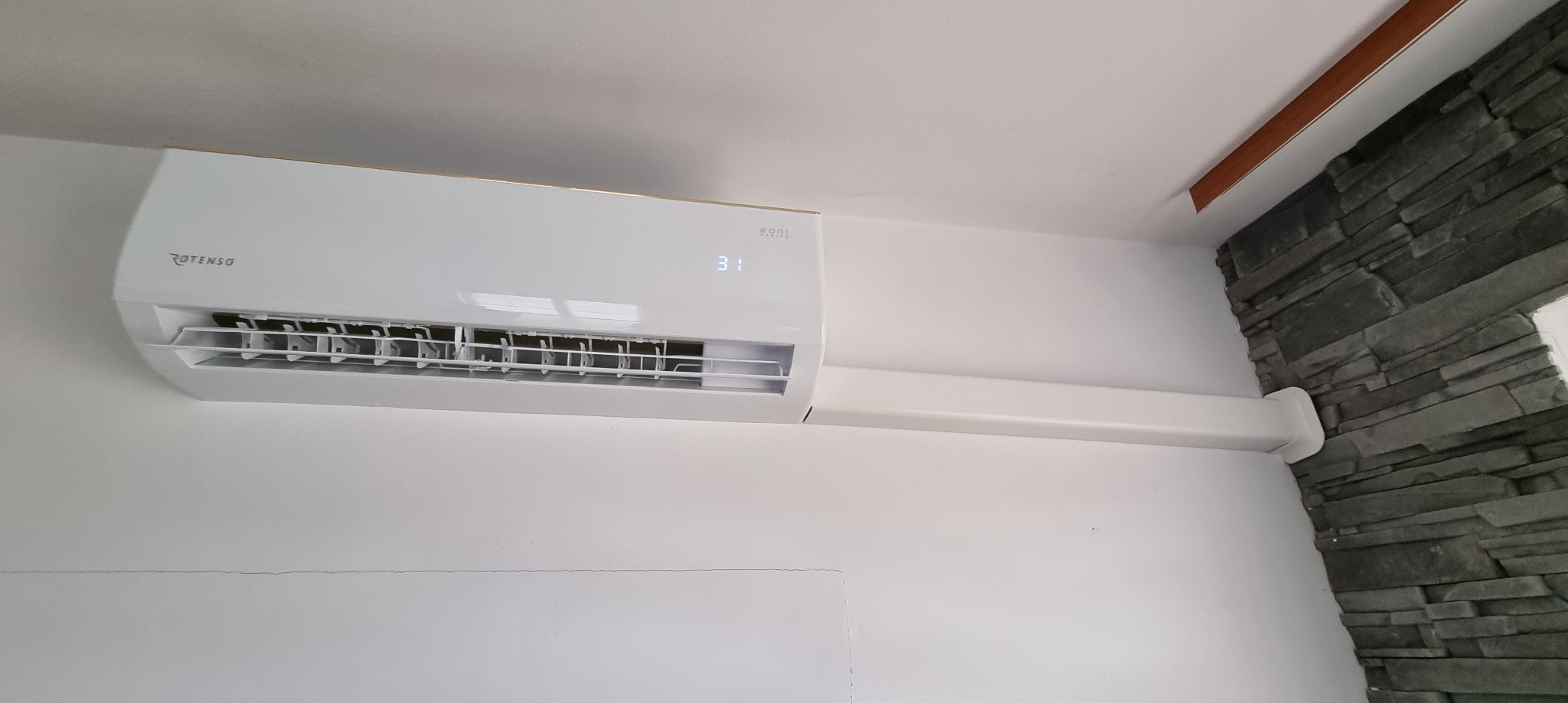 Biała klimatyzacja Rotenso z wyświetlaczem 31 stopni, zamontowana na białej ścianie obok dekoracyjnej ściany z szarego kamienia.