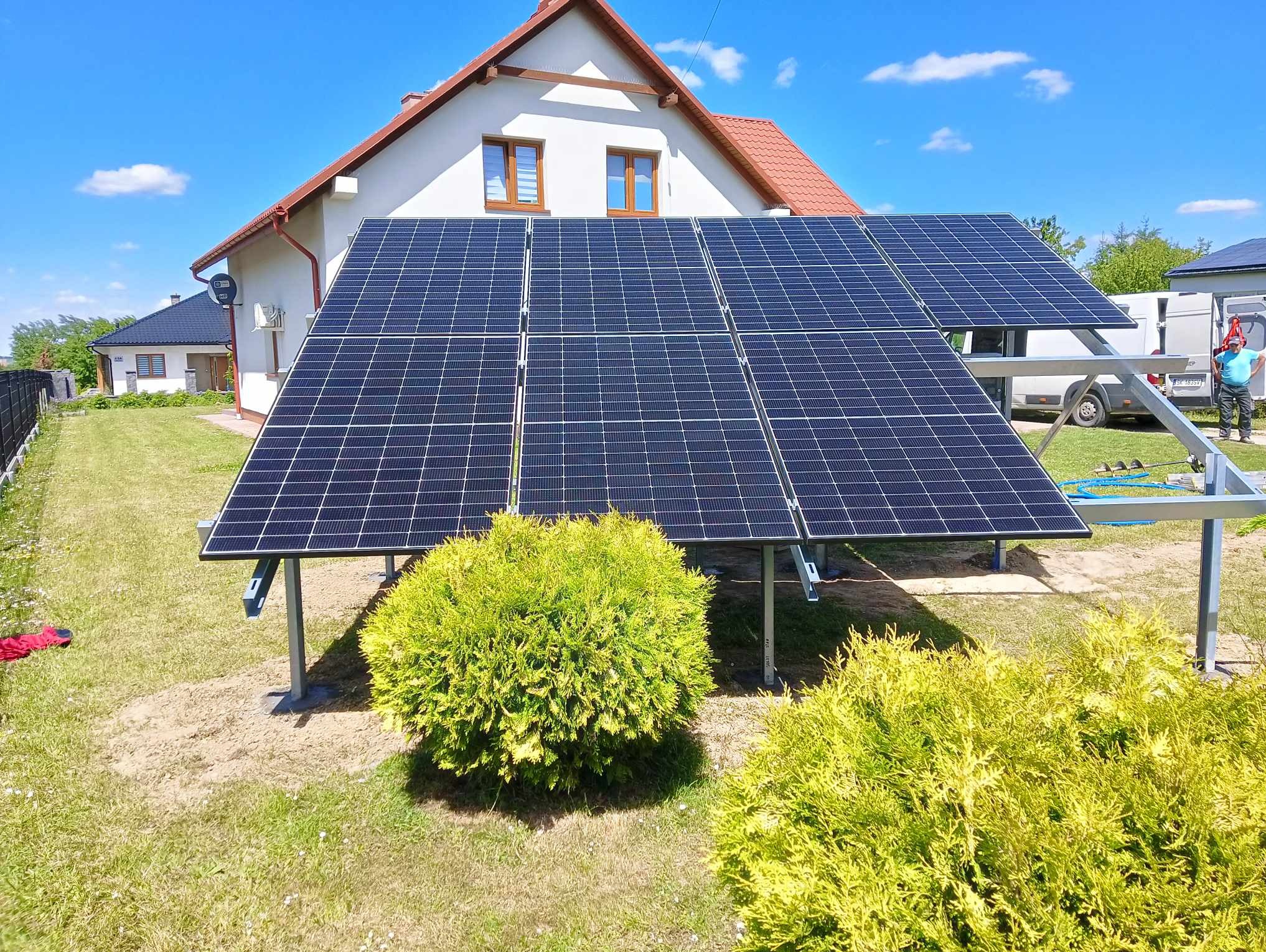 Instalacja paneli słonecznych na metalowej konstrukcji przed domem jednorodzinnym z czerwoną dachówką, widoczne krzewy i trawnik.