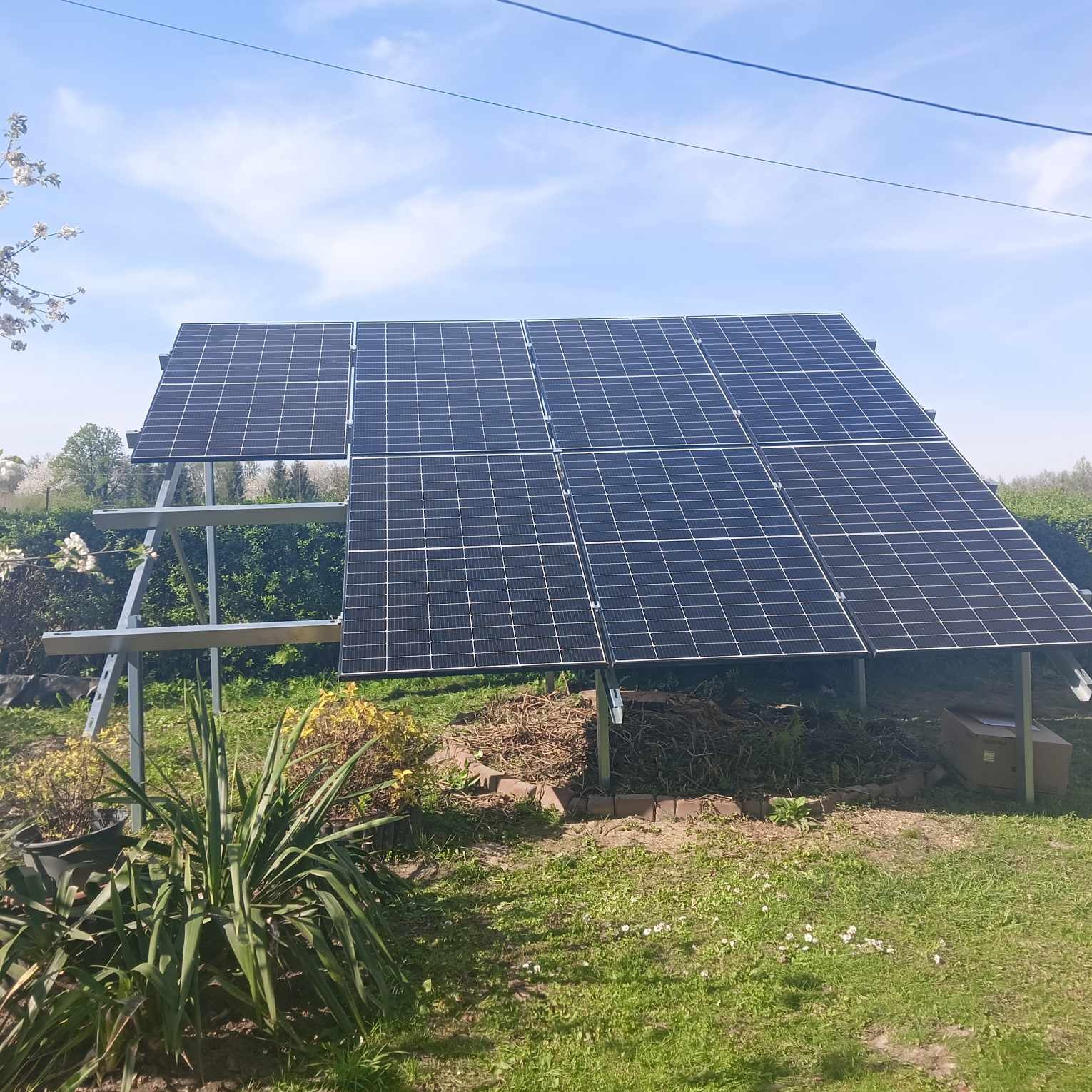 Instalacja naziemna paneli fotowoltaicznych na metalowej konstrukcji, widok z poziomu trawnika z roślinnością i kartonowym pudłem w tle, błękitne niebo.