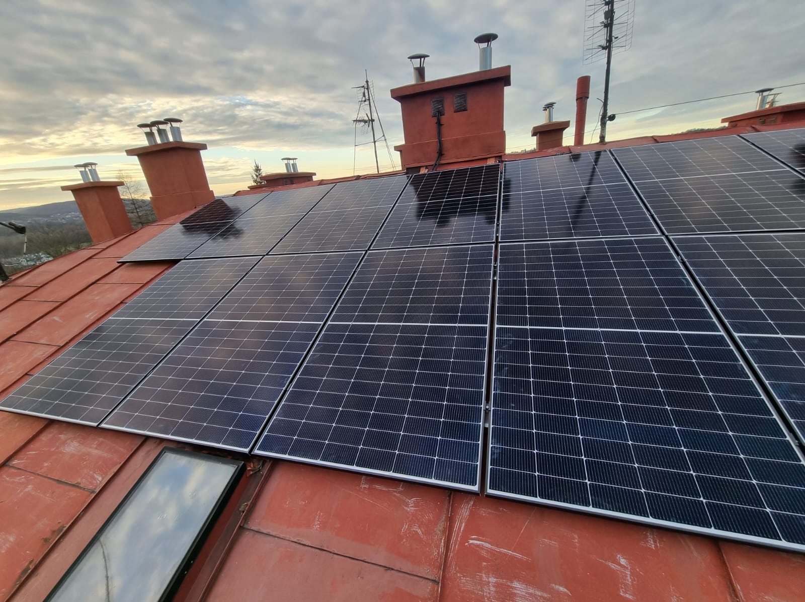 Panele słoneczne na czerwonym dachu z kominami i oknem dachowym, widok z góry pod pochmurnym niebem.