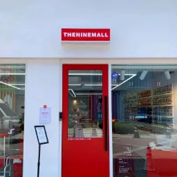 Wejście do sklepu THE NINEMALL z czerwoną ramą drzwi i szyldem nad wejściem, widoczne plakaty informacyjne i system alarmowy ADT.