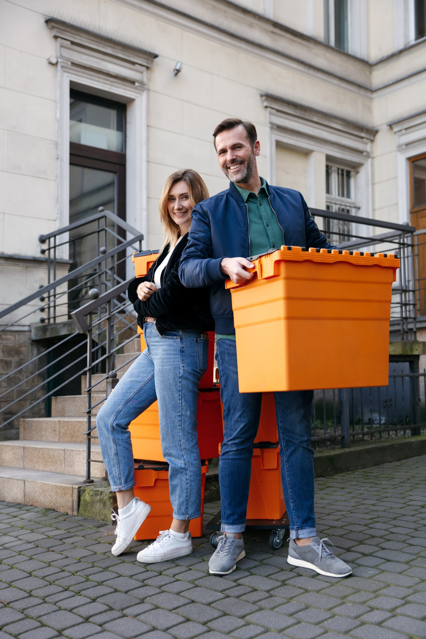 Uśmiechnięta para stoi przed budynkiem, trzymając pomarańczowe plastikowe pojemniki na kółkach, gotowa do transportu rzeczy.