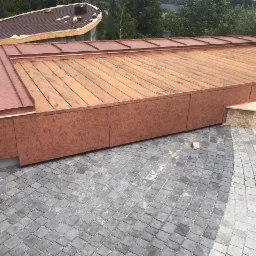 Wykończony taras z desek z obróbką blacharską w kolorze miedzi, widok z góry na szarą kostkę brukową, w tle budynek z dachem, na którym trwają prace.
