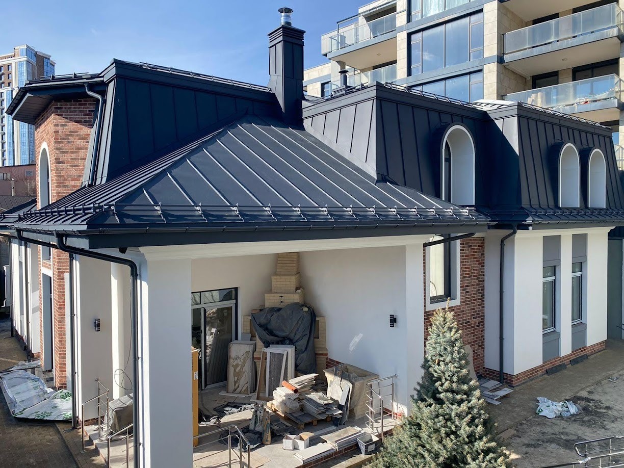 Nowoczesny dom z ciemnym dachem blaszanym i ceglanymi elementami, widok z góry na budynek w trakcie prac wykończeniowych, z widocznymi materiałami budowlanymi na tarasie i małą choinką w pobliżu.