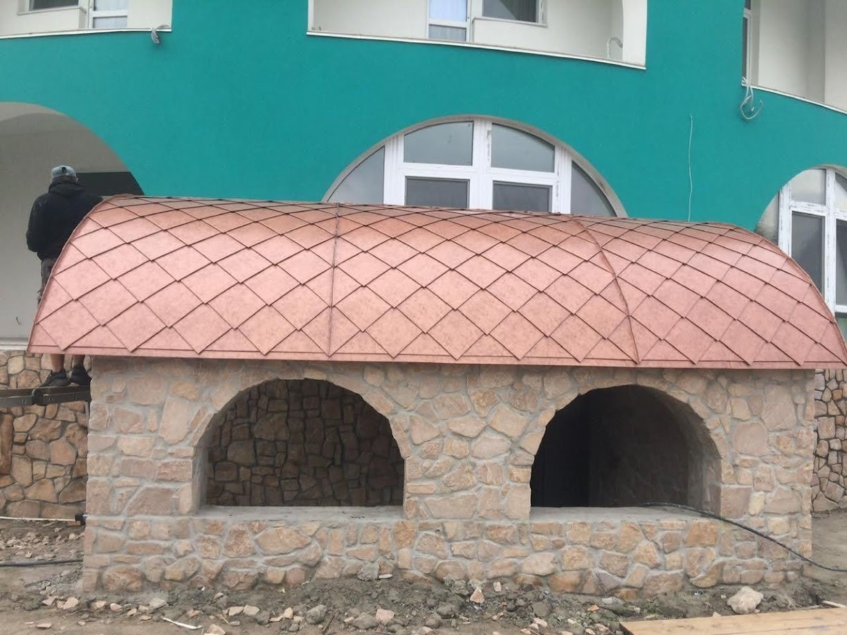 Kamienna konstrukcja z dwoma łukowatymi wnękami i miedzianym dachem łuskowym w Warszawie, widoczny pracownik na rusztowaniu w tle.