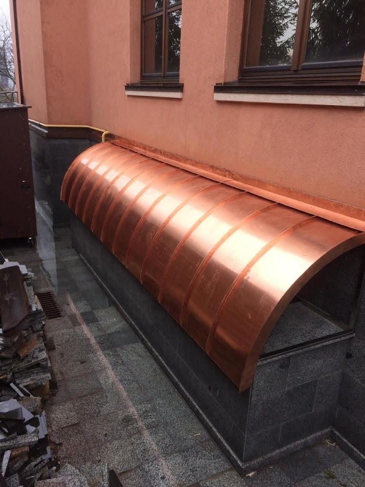 Miedziane zadaszenie nad zejściem do piwnicy, wykonane z segmentów, przymocowane do ściany budynku z oknami, widoczne kamienne wykończenie i stos kamieni.