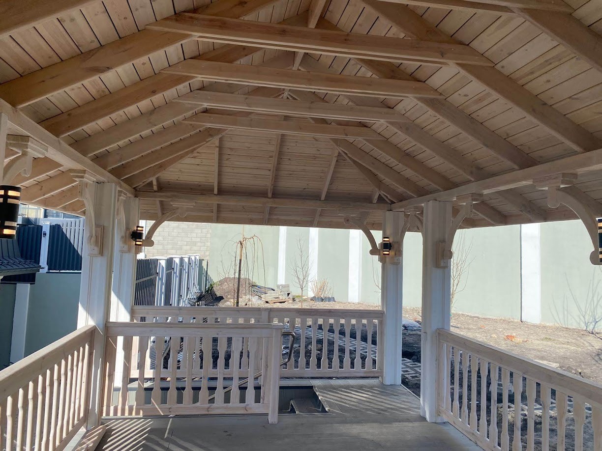 Surowa drewniana konstrukcja altany z balustradą, widok od wewnątrz, z zamontowanymi lampami ściennymi, teren wokół w trakcie przygotowania.