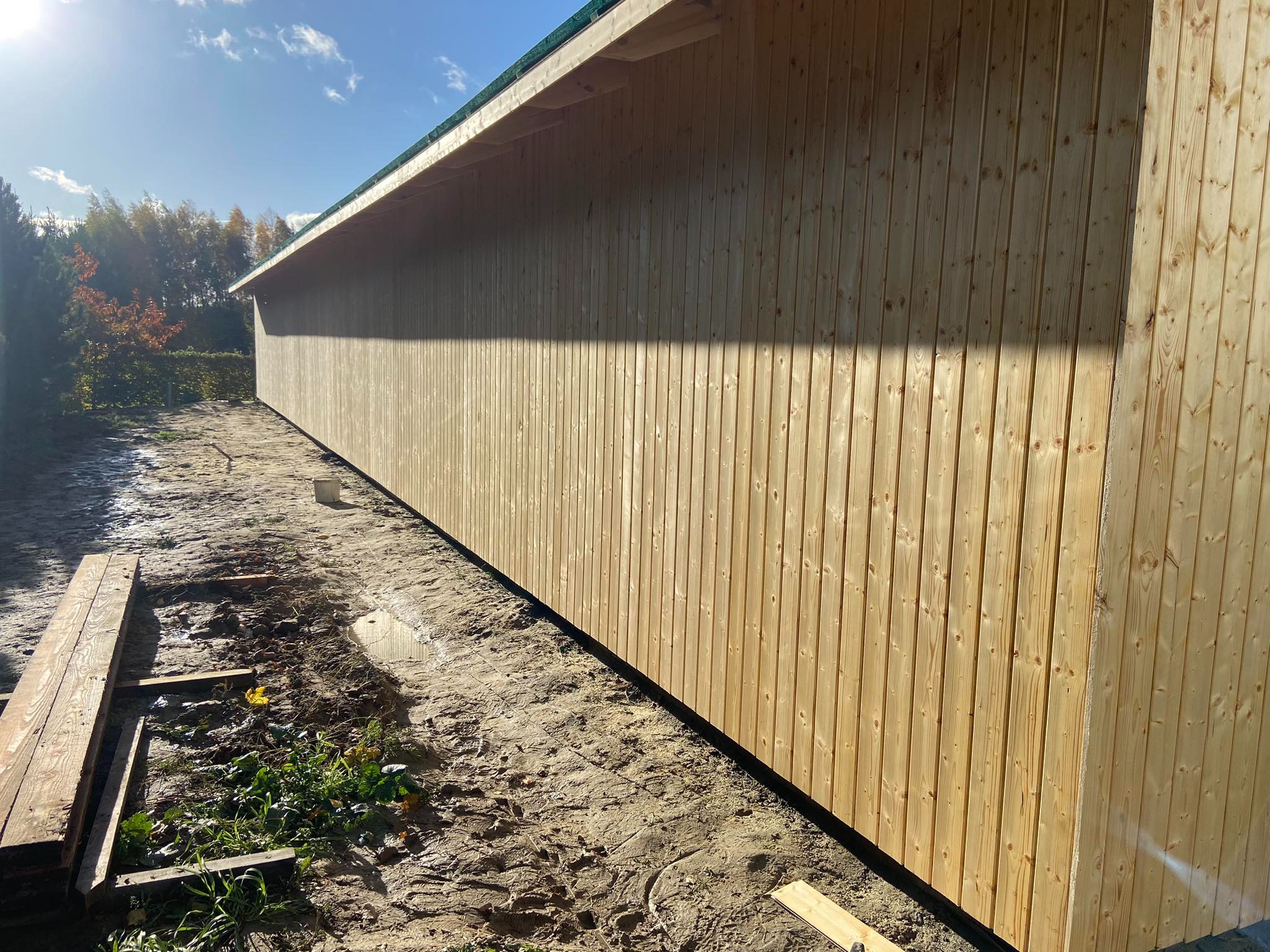 Ściana zewnętrzna budynku w trakcie budowy, obłożona jasnymi, pionowymi deskami boazeryjnymi, widoczne niedoskonałości terenu i składowane deski.