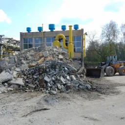 Sterta gruzu po rozbiórce budynku przemysłowego z żółtymi rurami wentylacyjnymi, w tle żółta ładowarka kołowa i ciężarówka.