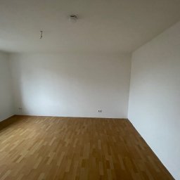 ZACHIBAU usługi ogólnobudowlane - Pusty pokój z drewnianą podłogą, świeżo pomalowanymi białymi ścianami, grzejnikiem pod oknem i czujnikiem dymu na suficie, gotowy do umeblowania.
