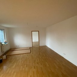 ZACHIBAU usługi ogólnobudowlane - Pusty pokój po remoncie z drewnianą podłogą, białymi ścianami, grzejnikiem pod oknem, dwoma panelami podłogowymi i dwoma grzejnikami elektrycznymi.