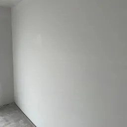 Świeżo wyszpachlowana ściana w rogu pomieszczenia, widoczne niedoskonałości na gładkiej powierzchni i szary betonowy podkład pod podłogę.