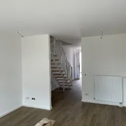 Jasne, przestronne wnętrze w trakcie wykańczania: świeżo pomalowane białe ściany, drewniane panele podłogowe, schody z białymi barierkami, grzejnik, wystające kable z sufitu i zwój paneli...