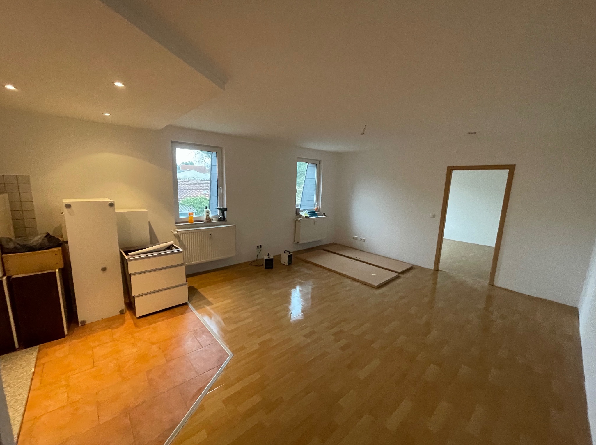 Przestronne wnętrze w trakcie przygotowań do remontu; widoczne odsłonięte panele podłogowe, grzejniki pod oknami, oraz przygotowane meble do zabezpieczenia.