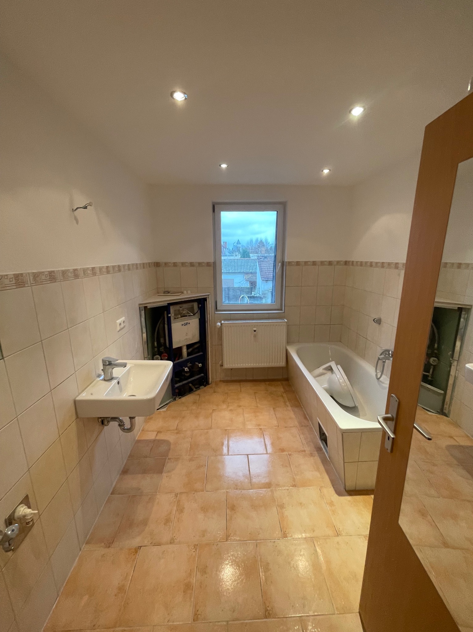 Łazienka w trakcie remontu: widoczna wanna z zabezpieczeniem, umywalka, odsłonięta instalacja wodna i grzejnik pod oknem.