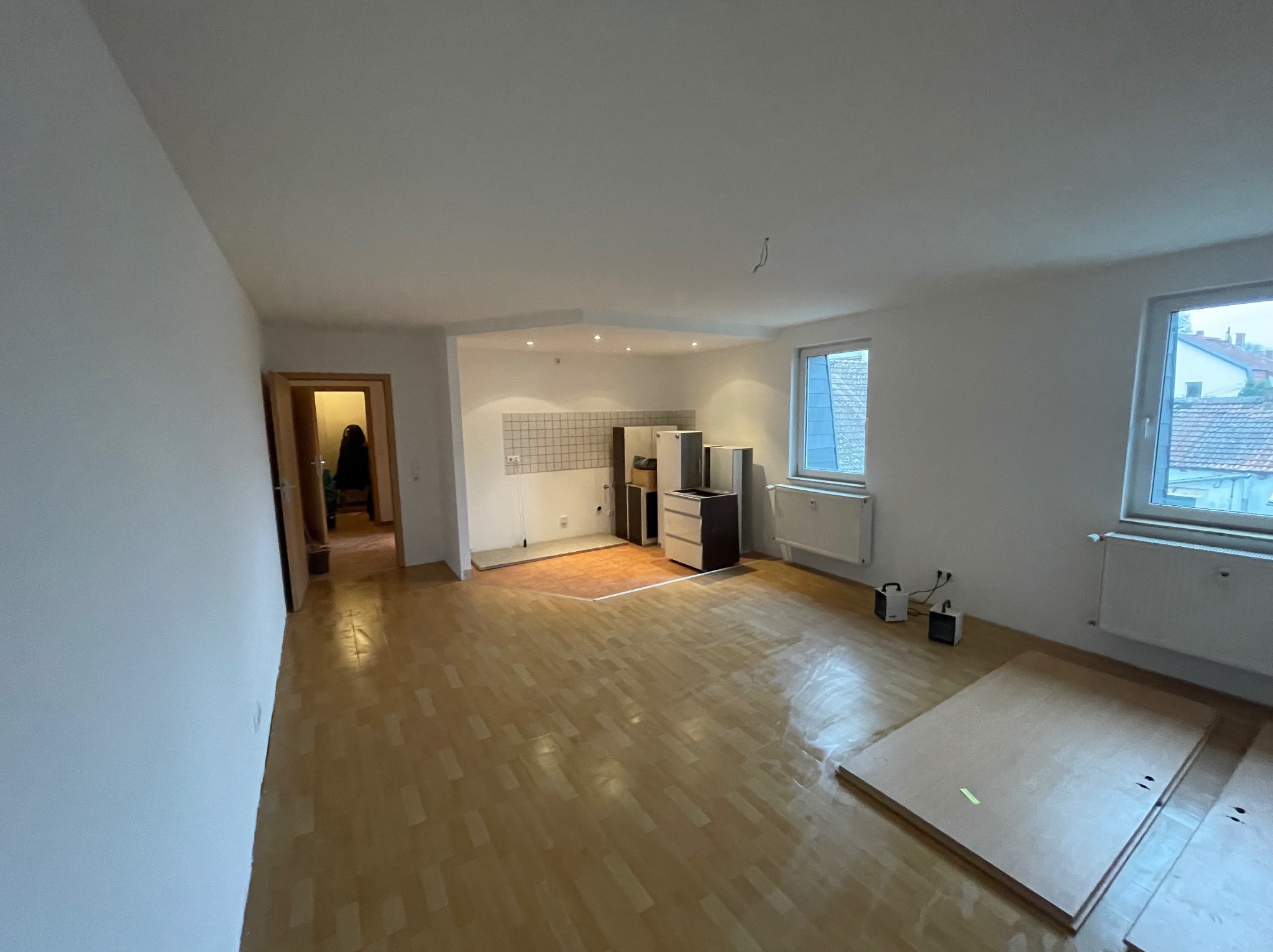 Wnętrze pustego pokoju z parkietem, widoczne ściany przygotowane do malowania, fragment kuchni z zabudową oraz okna z widokiem na zewnątrz.