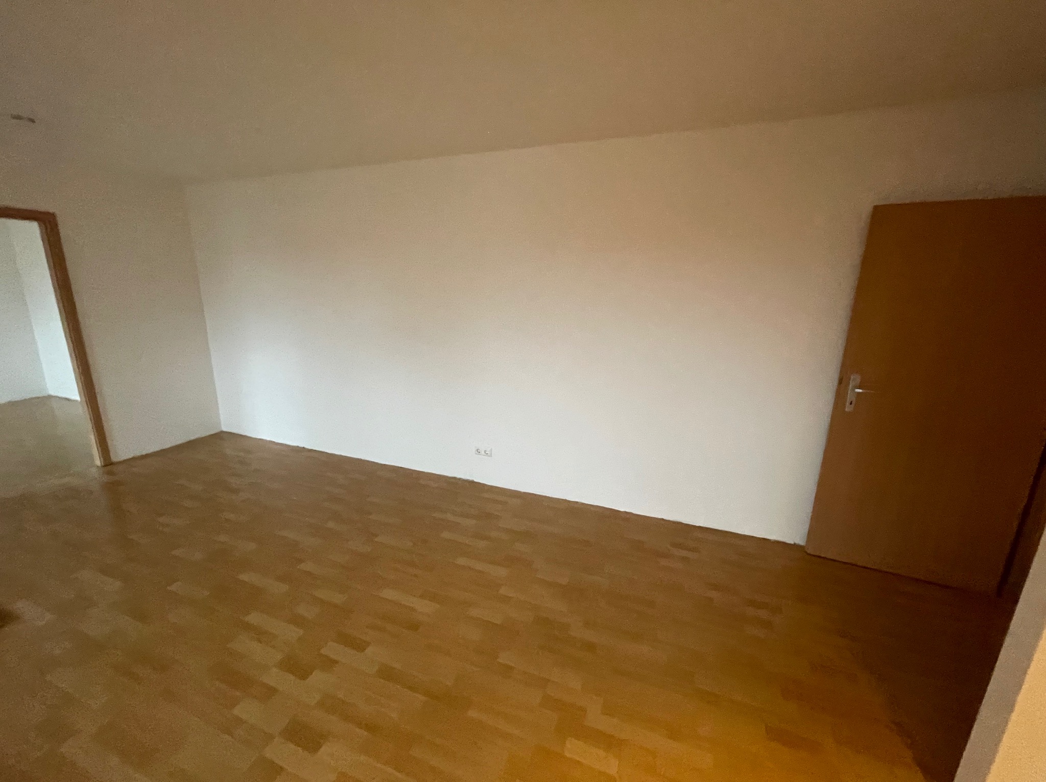 Pusty pokój z parkietem ułożonym w jodełkę, świeżo pomalowanymi na biało ścianami i brązowymi drzwiami po prawej stronie, przygotowany do dalszych prac wykończeniowych.