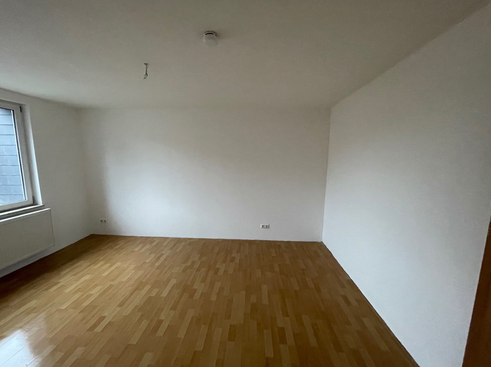 Pusty pokój z drewnianą podłogą, świeżo pomalowanymi białymi ścianami, grzejnikiem pod oknem i czujnikiem dymu na suficie, gotowy do umeblowania.