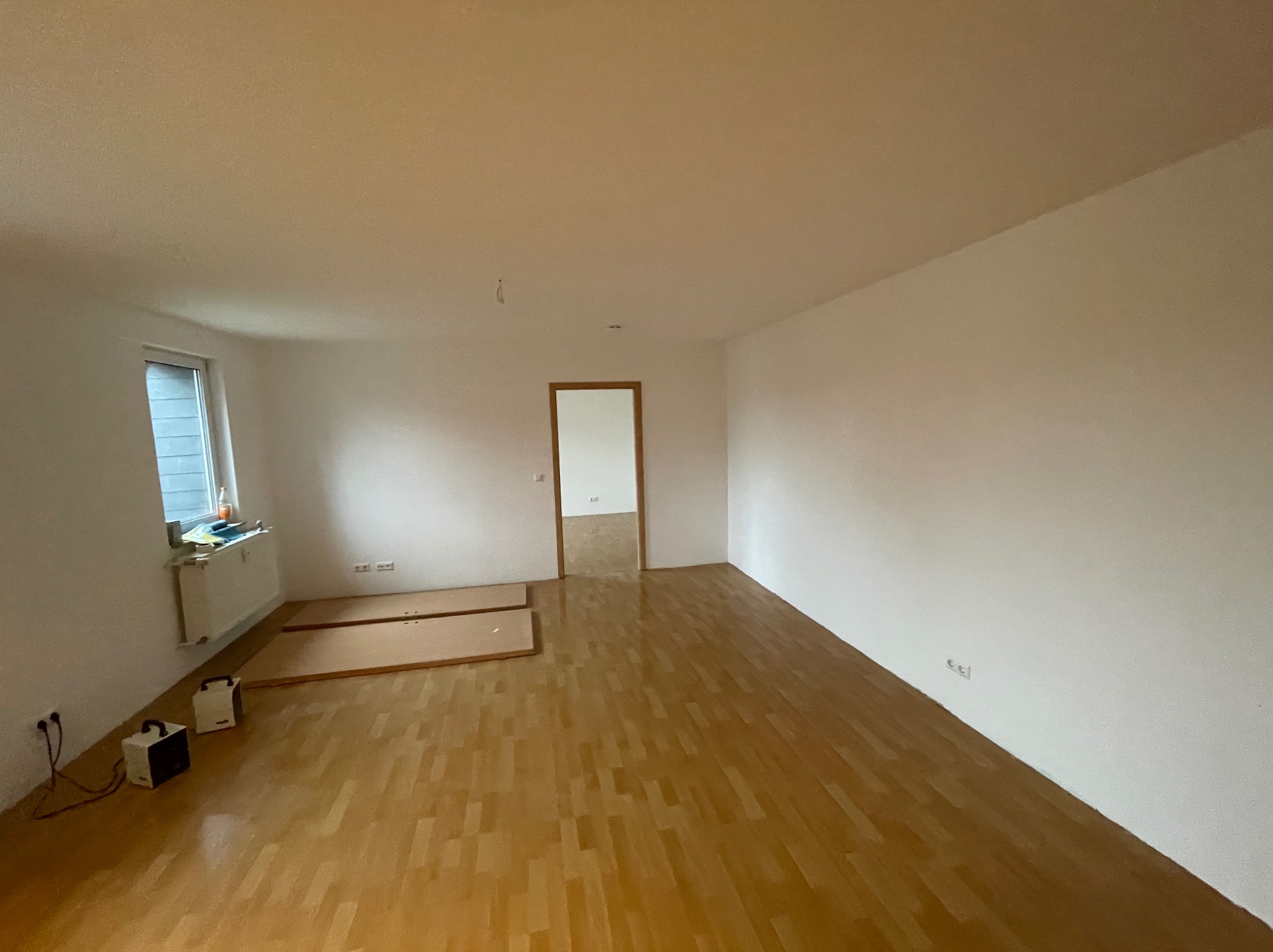 Pusty pokój po remoncie z drewnianą podłogą, białymi ścianami, grzejnikiem pod oknem, dwoma panelami podłogowymi i dwoma grzejnikami elektrycznymi.