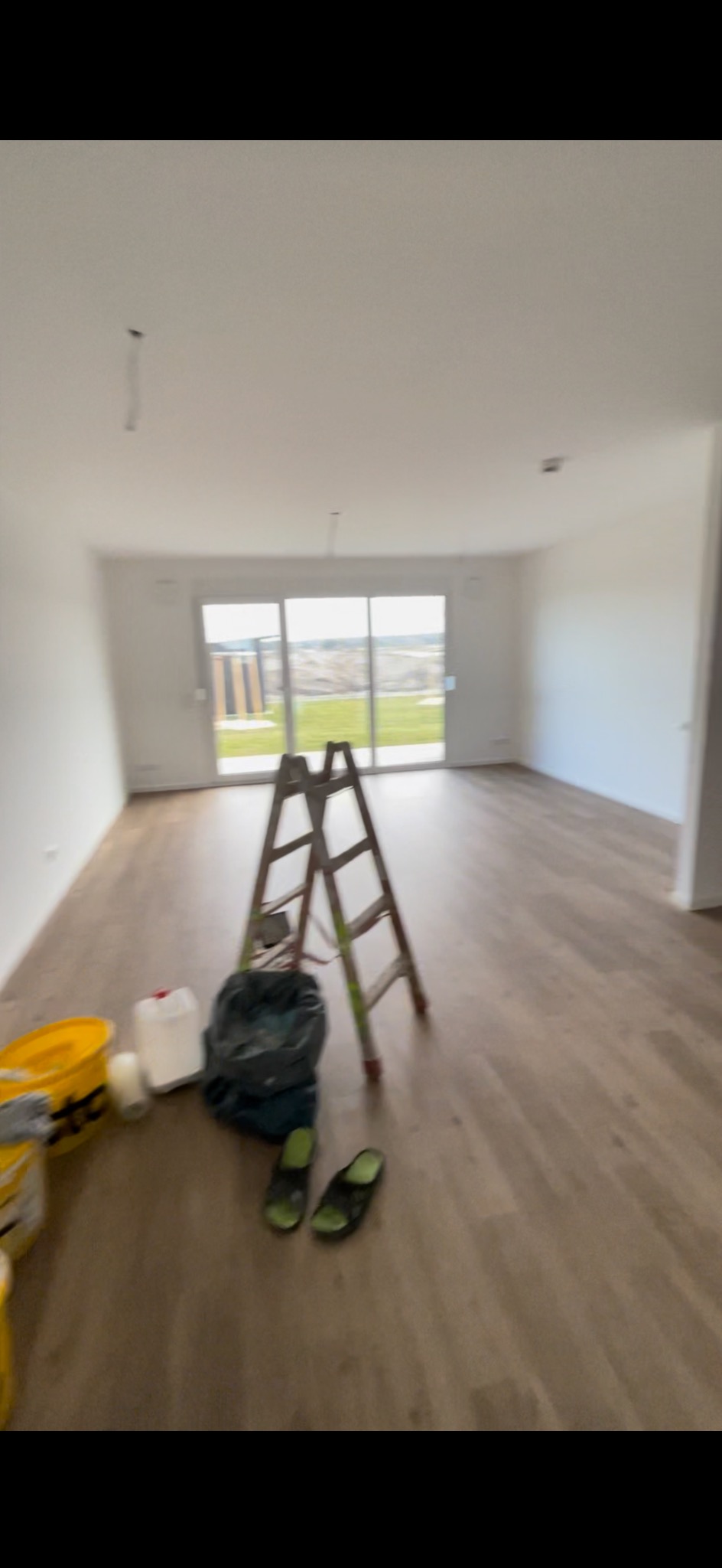 Wnętrze pokoju z drewnianą podłogą w trakcie prac, widoczna drabina, wiadro z farbą, pojemnik i klapki na podłodze, duże okno balkonowe w tle.