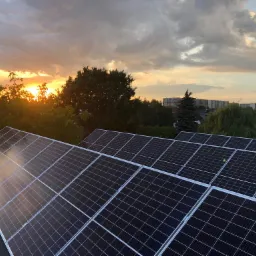 Panele fotowoltaiczne na dachu domu widziane z góry, z zachodzącym słońcem i drzewami w tle.