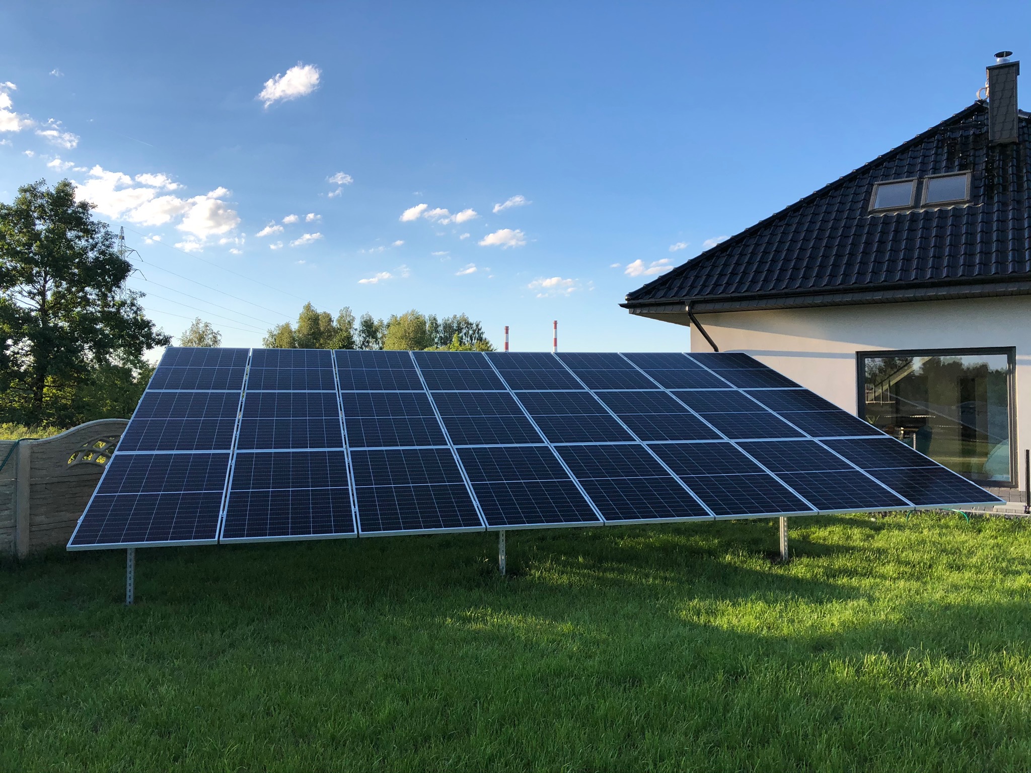 Panel słoneczny ustawiony na trawniku przed domem jednorodzinnym z ciemnym dachem i oknami, widoczne niebo z chmurami i drzewa w tle. Konstrukcja wsporcza paneli wykonana z metalowych profili.
