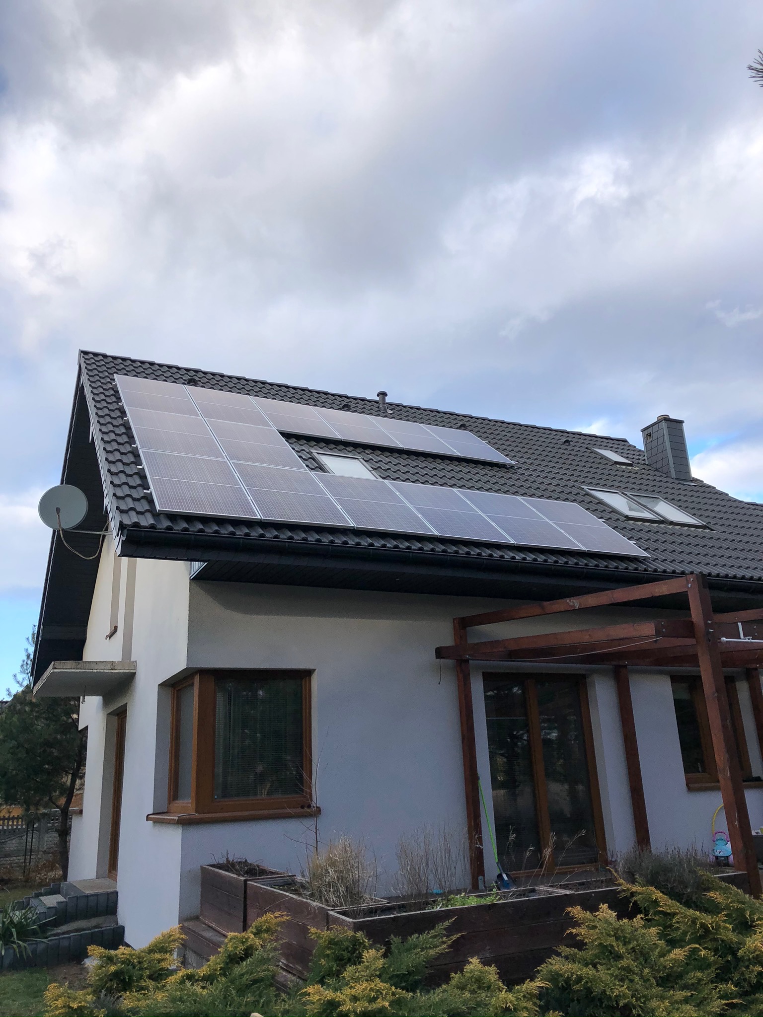 Dom jednorodzinny z zamontowanymi panelami słonecznymi na dachu pokrytym ciemną dachówką, widoczne okna dachowe, antena satelitarna oraz pergola.