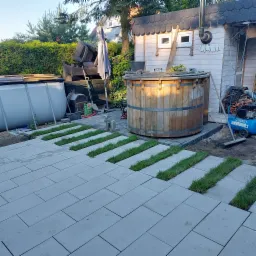 Ułożona szara kostka brukowa z pasami świeżo posadzonej trawy, drewniana beczka z wodą i budynek gospodarczy w tle.