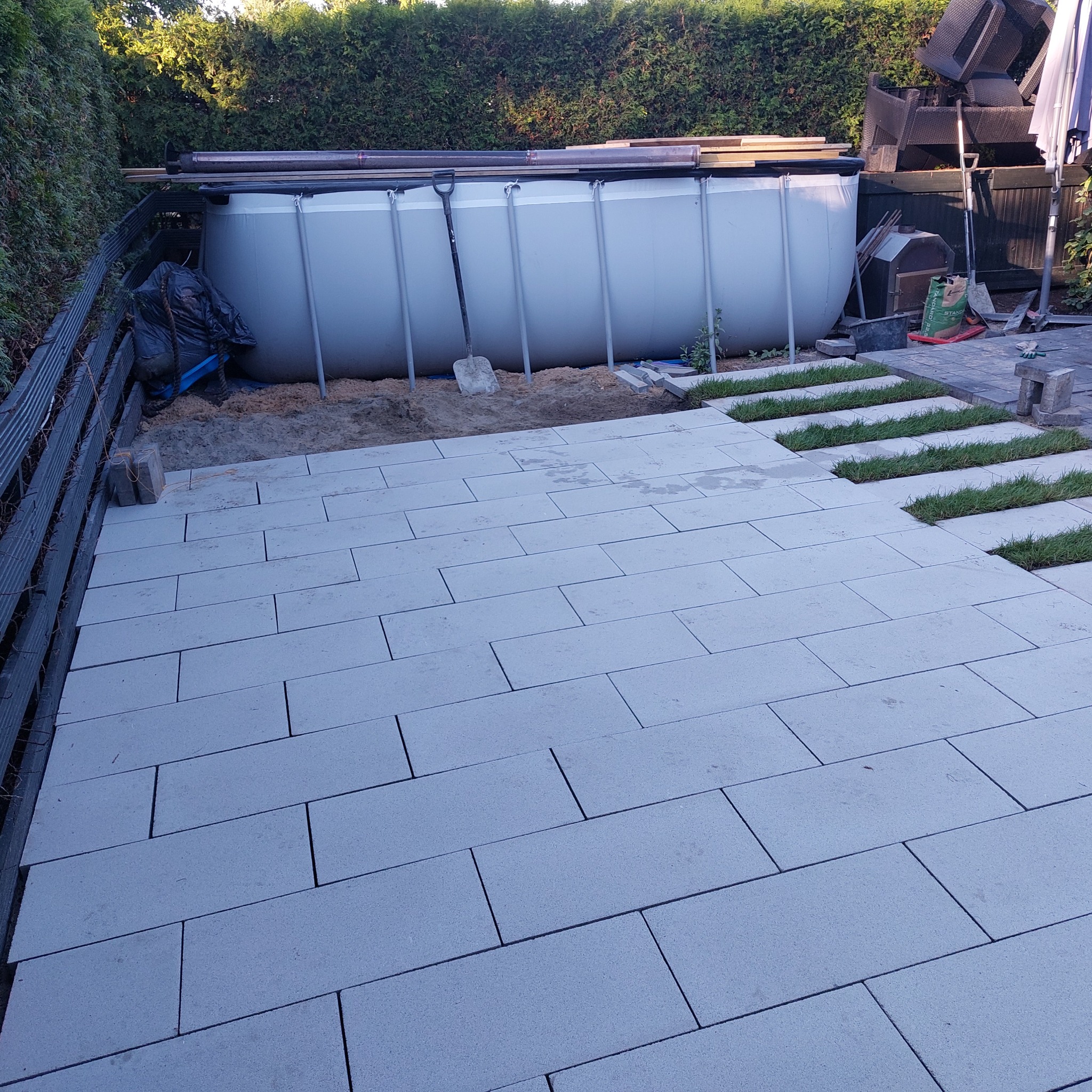 Jasnoszare prostokątne płyty ułożone na podwórku z fragmentami trawy, w tle basen ogrodowy i narzędzia.