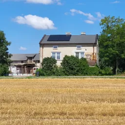 Dom jednorodzinny z zainstalowanymi panelami fotowoltaicznymi na dachu, widok z pola po żniwach w słoneczny dzień.