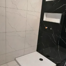 Kabina prysznicowa z białym brodzikiem i płytkami imitującymi marmur w kolorze białym i czarnym, z wnękami w ścianie wyłożonymi płytkami.