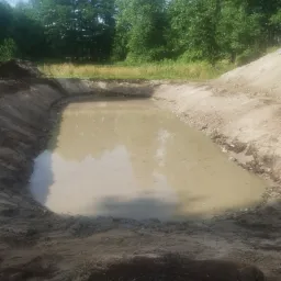Wykopany staw wypełniony wodą, otoczony ziemią i zielenią drzew w tle.