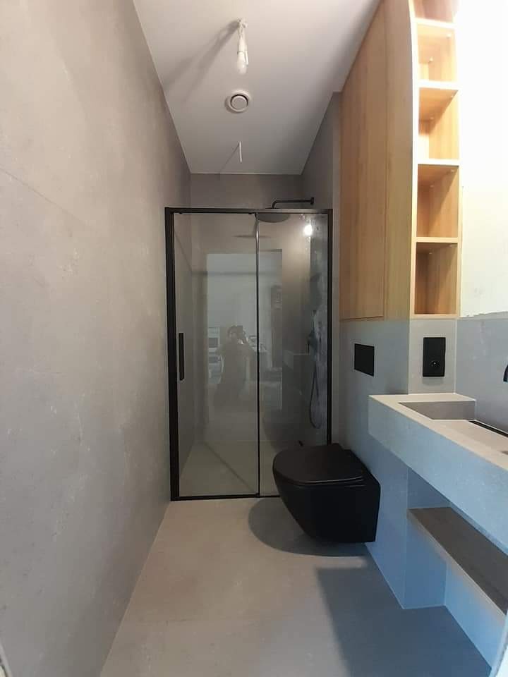 Nowoczesna łazienka z kabiną prysznicową w czarnej ramie, wiszącą czarną toaletą i umywalką z szarego konglomeratu na tle ściany w odcieniach szarości, oświetlona wiszącą żarówką.