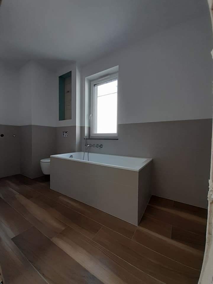 Wnętrze łazienki w trakcie wykańczania: zabudowana wanna, drewnopodobne płytki na podłodze, ściany pomalowane na szaro i biało, widoczne przyłącza wodne, brak fug w płytkach wanny.